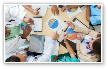 Hospital administrators and medical professionals review pie charts and graphs during staff meeting  Professionals are gathered around a round table in a board room  Photo taken from high angle viewpoint 